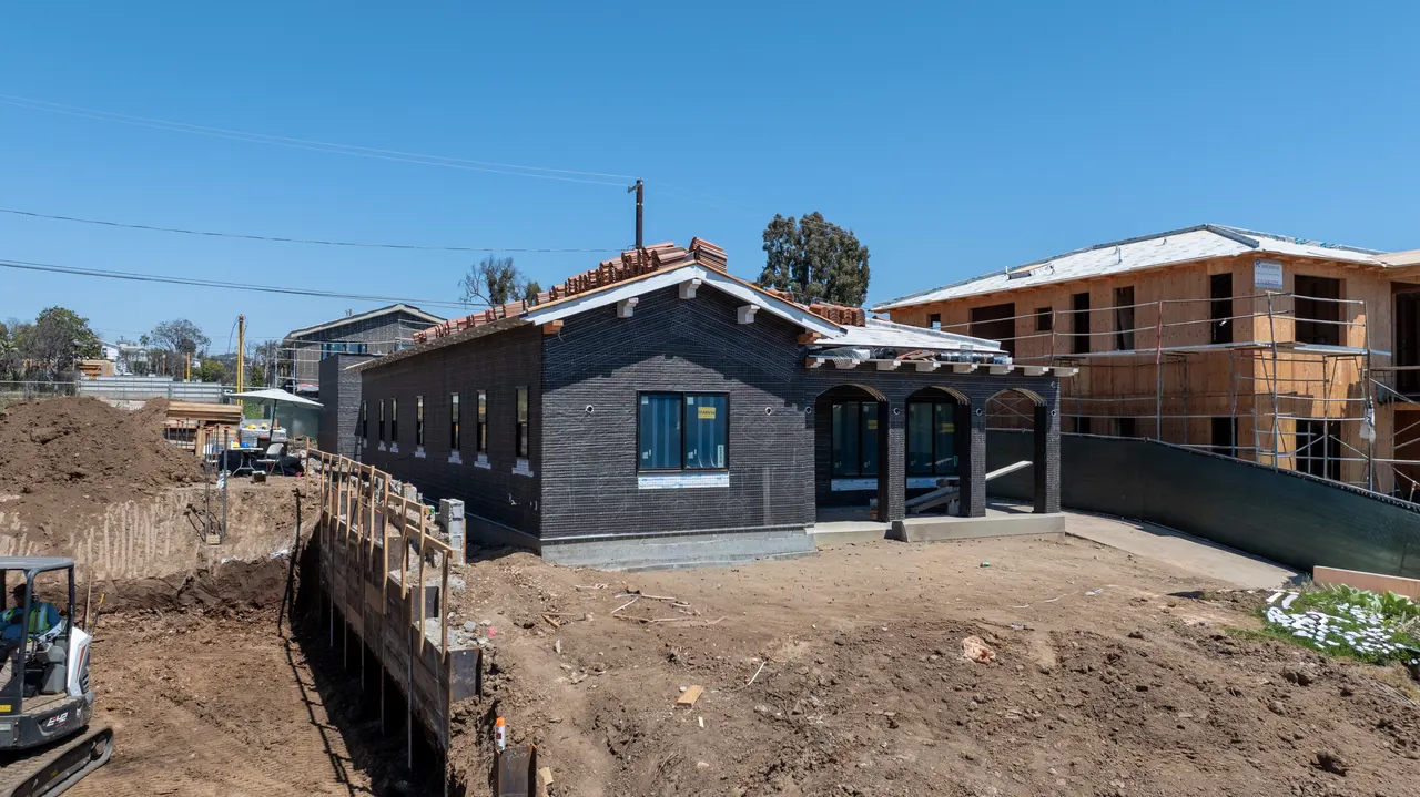 Aerial view of a home nearing completion with dark wire-lath walls and a red clay tile roof partway through installation, bare dirt yard, and neighboring homes under construction