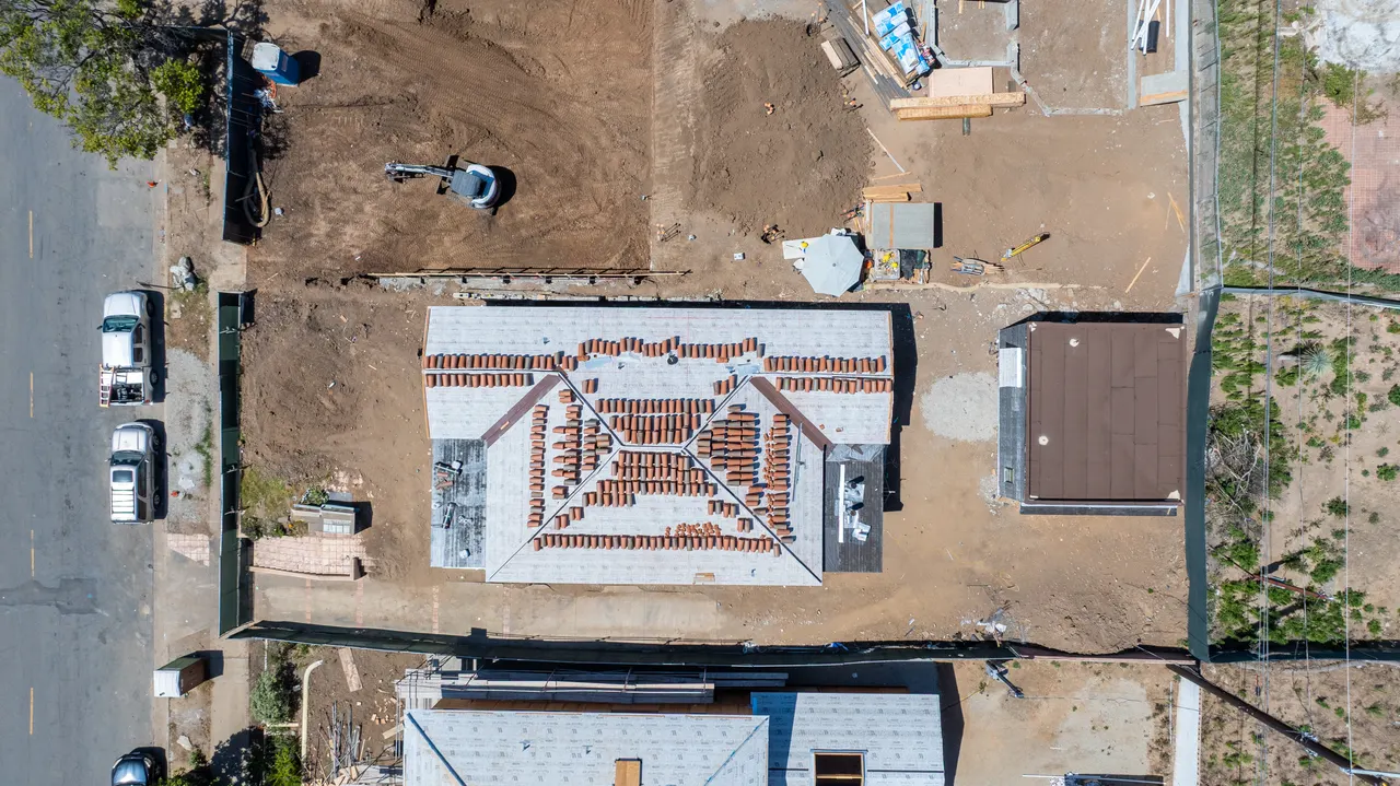 Aerial view of clay roof tiles staged and being laid across the ridges and hips of the roof, with a detached dark-clad accessory structure alongside