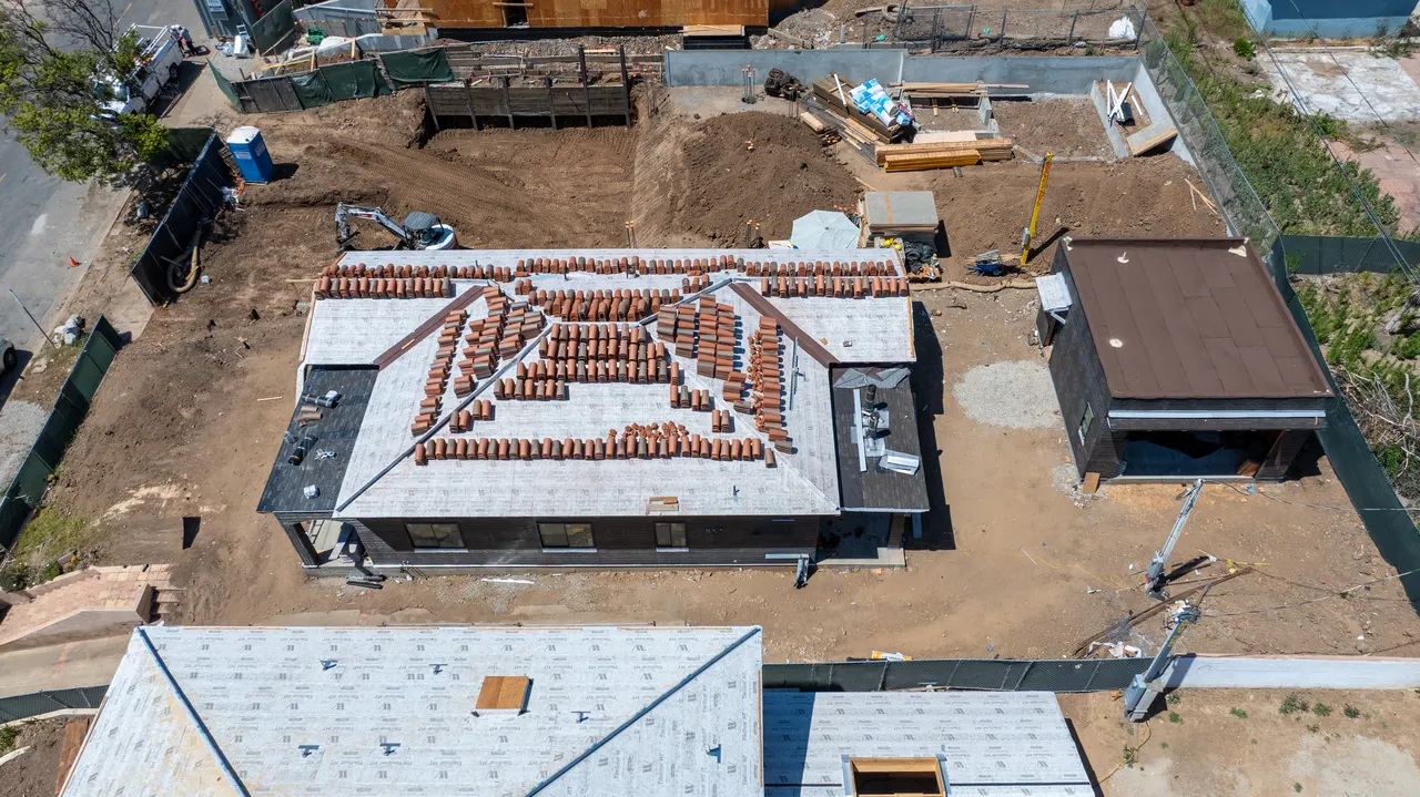 Aerial view of the roof mid-tile-installation with roofing materials staged on top, a detached accessory structure, and dirt yard below