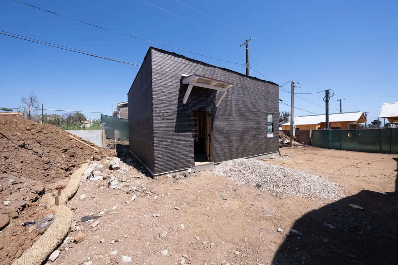 Detached accessory structure wrapped in dark wire lath with a wood door header, surrounded by dirt and construction debris