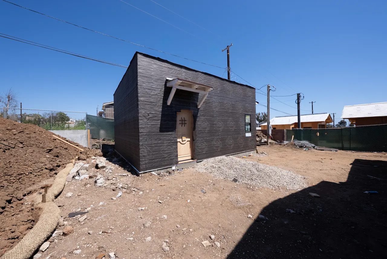 Detached accessory structure with dark wire-lath siding and a wood entry door installed, dirt yard in front