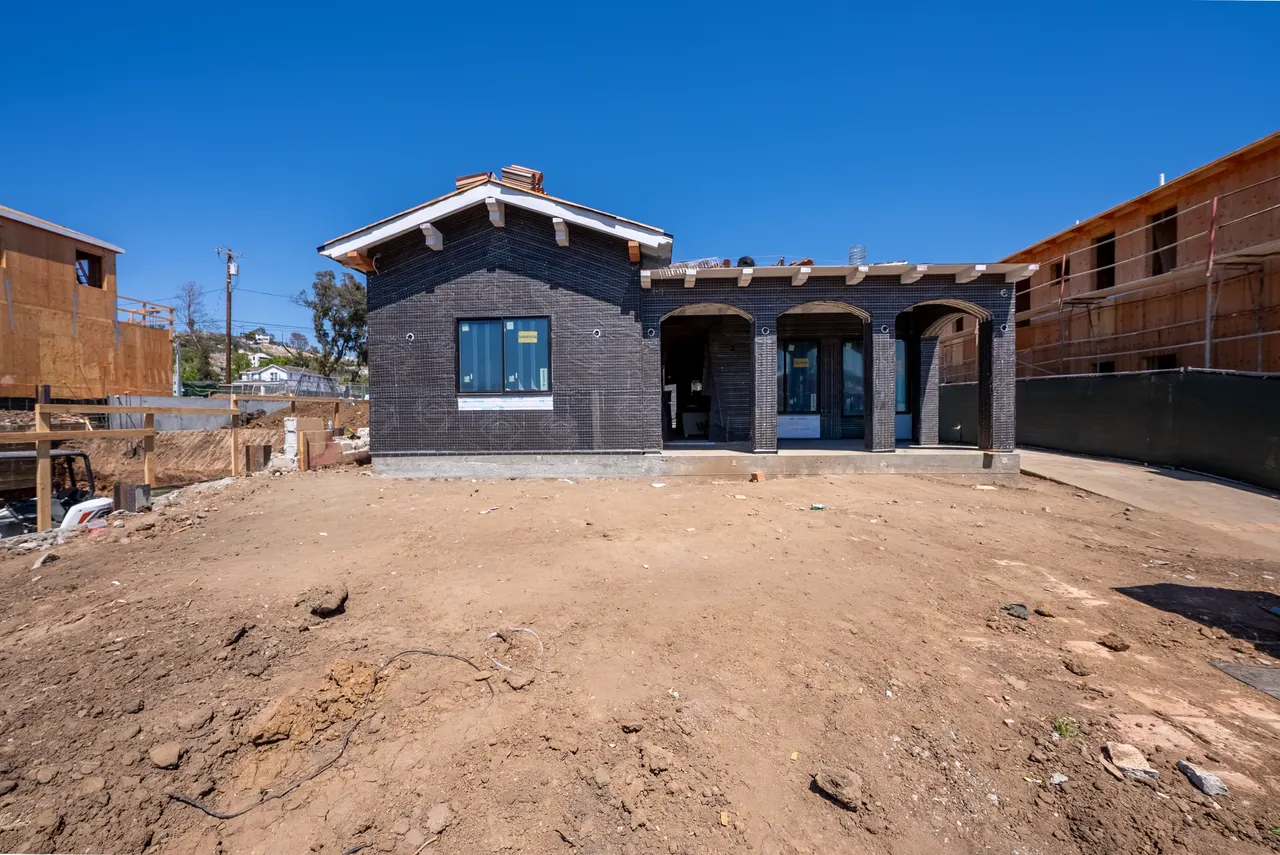 Front elevation of the home with dark wire-lath cladding, a gabled roof with clay tile ridge, an arched covered entry, and a bare dirt yard