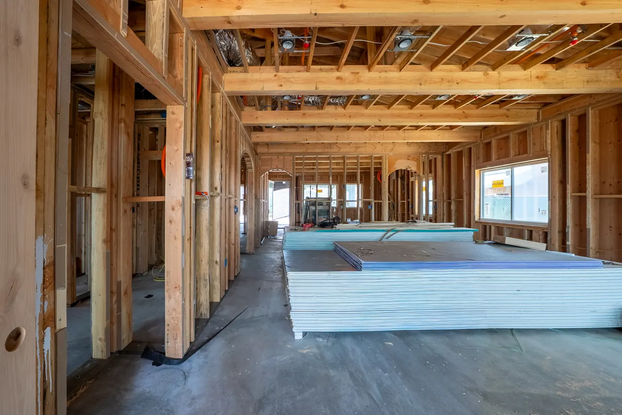 Framed great room with exposed wood ceiling beams, an arched doorway, stacked drywall sheets on the floor, and a wall of windows