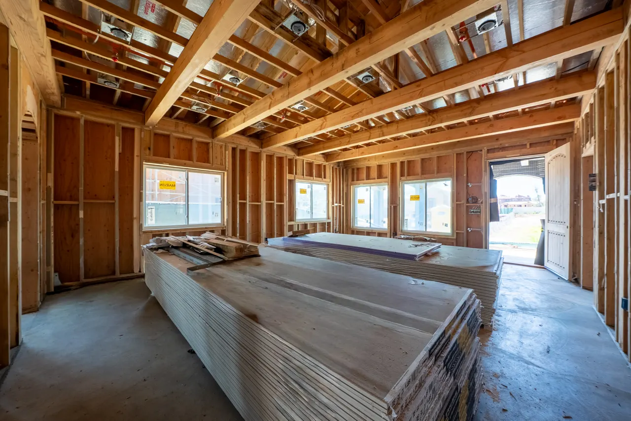 Framed great room with a vaulted wood beam ceiling, a bank of windows, an arched wood door open to the yard, and stacked drywall on the floor