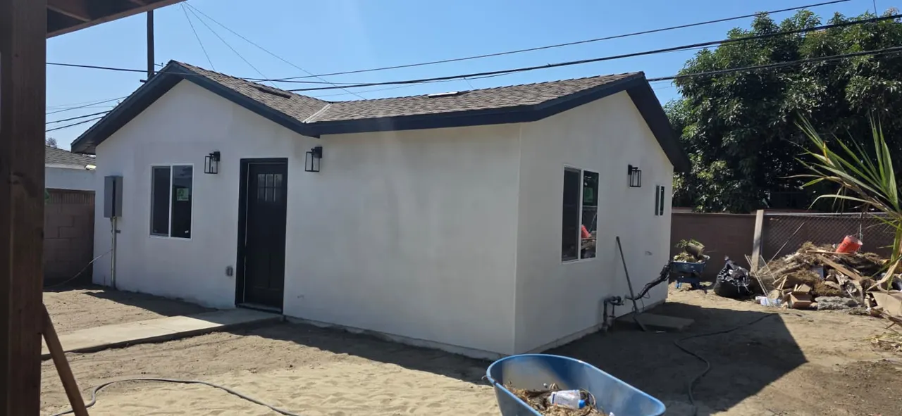 Finished white stucco detached ADU, corner view showing the black front door, two windows, wall sconces, and gray shingle roof against a blue sky