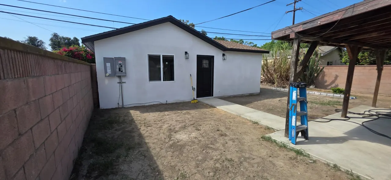 Front elevation of the white stucco ADU with a black door, window, wall sconces, exterior electrical panel, and a concrete walkway leading to a wood-framed patio cover alongside