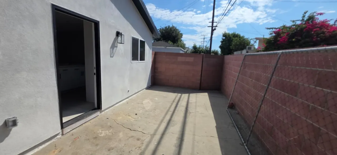 Side-yard walkway along the ADU with an open black door revealing kitchen cabinets inside, a concrete block wall, chain-link fencing, and a flowering shrub