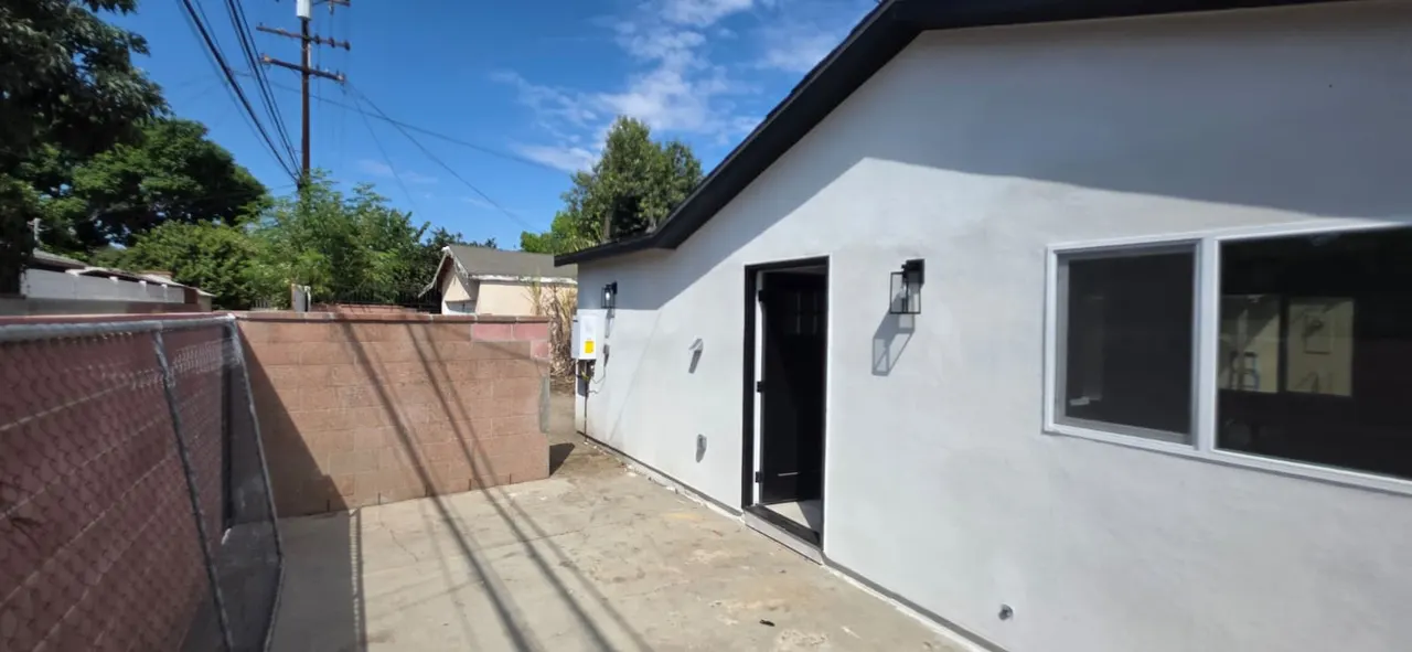 Corner of the white stucco ADU with an open black door, wall sconce, red concrete block wall, and chain-link fence along the side yard