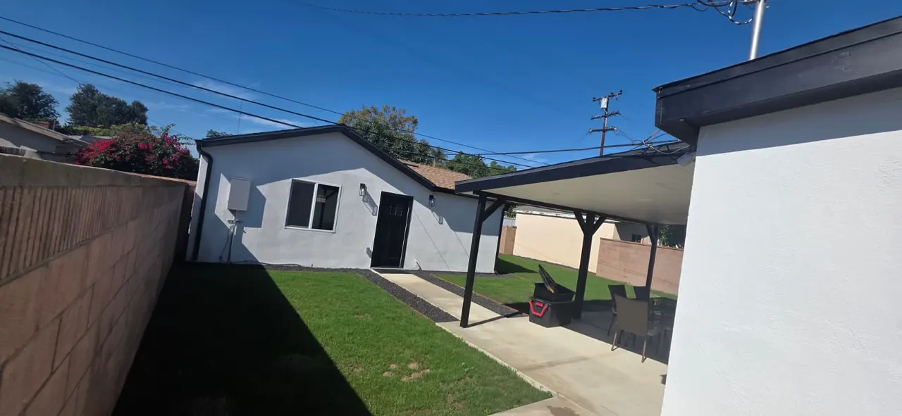 White stucco ADU exterior with turf lawn, black door and window, and a covered patio with outdoor furniture, viewed from a different angle