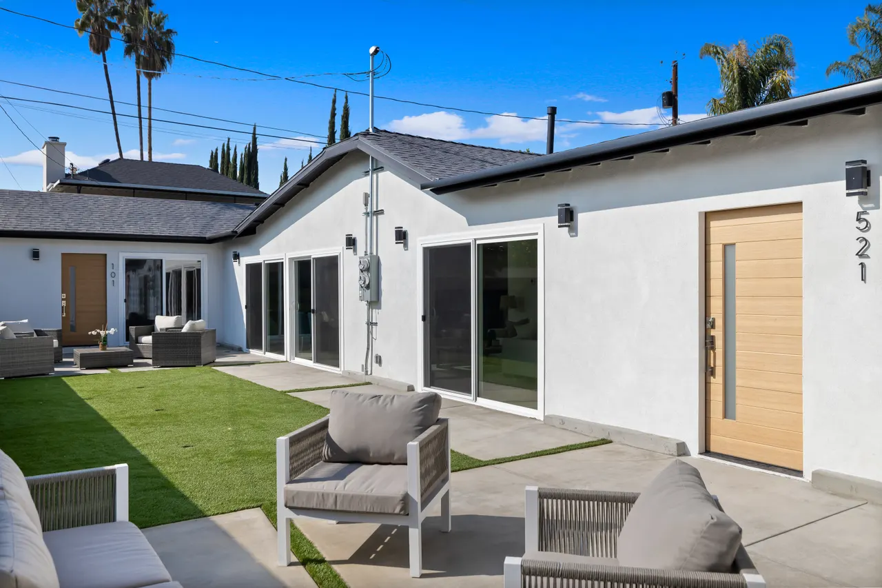 Rear exterior showing the full width of the home, a turf lawn strip along a stucco privacy wall, outdoor sofa and lounge chairs on a concrete patio, and palm trees under a blue sky