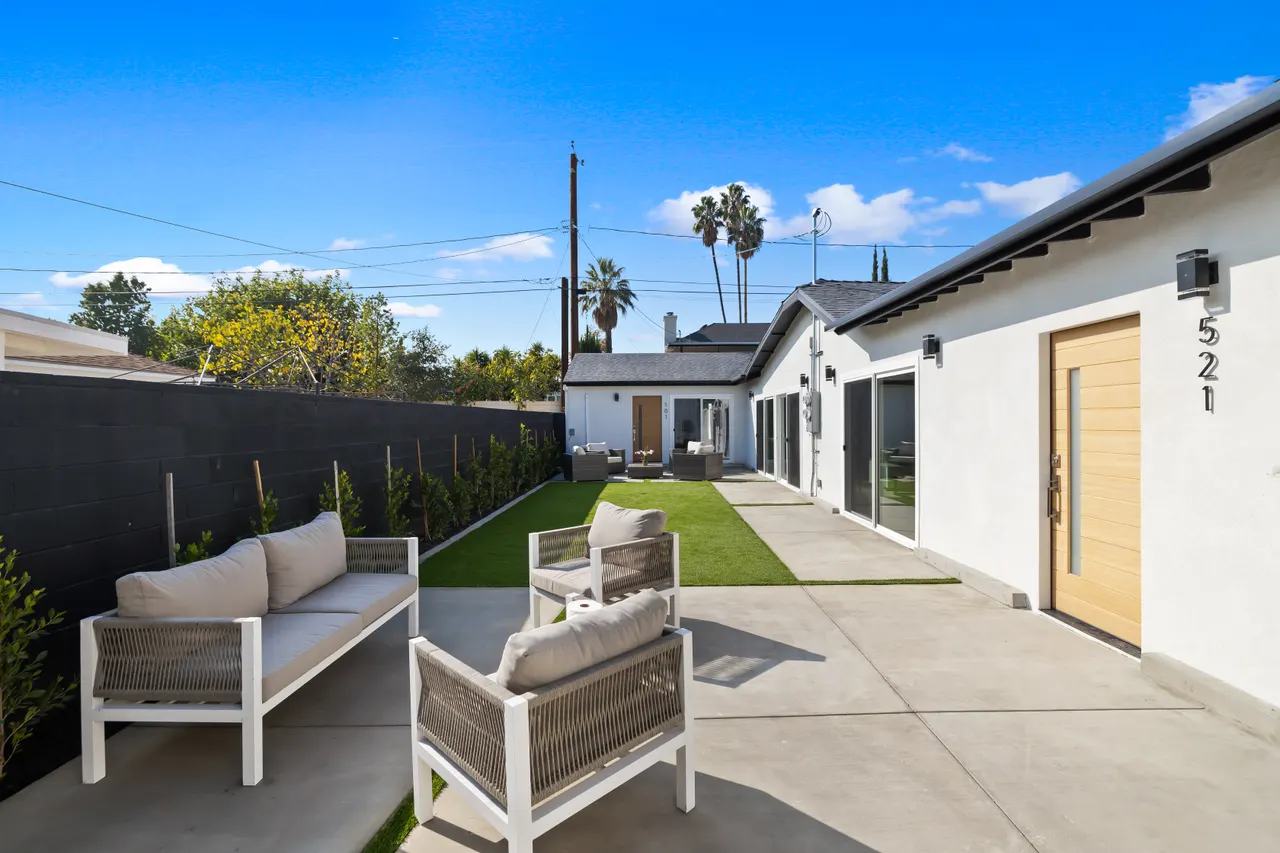 Side-yard patio nook with a narrow turf strip along a tall fence, an outdoor sofa and armchair with cushions, and a small side table