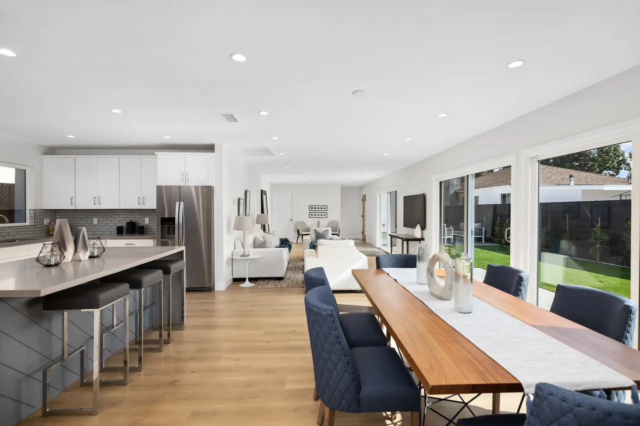 Dining area within the open great room, a long wood table with navy upholstered chairs, and the kitchen island with bar seating beyond