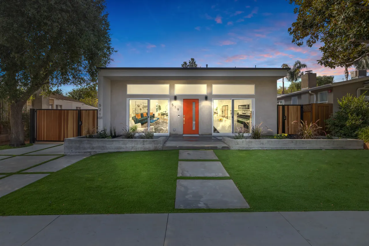 Twilight exterior of the home with the orange front door and windows glowing warm light against a dusk sky, landscape lighting along the lawn and pavers