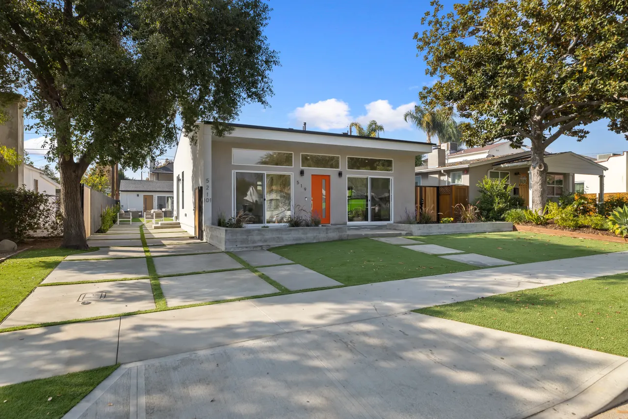 Front exterior viewed from the driveway, mature trees framing the house with the orange door visible at the end of the paver walkway