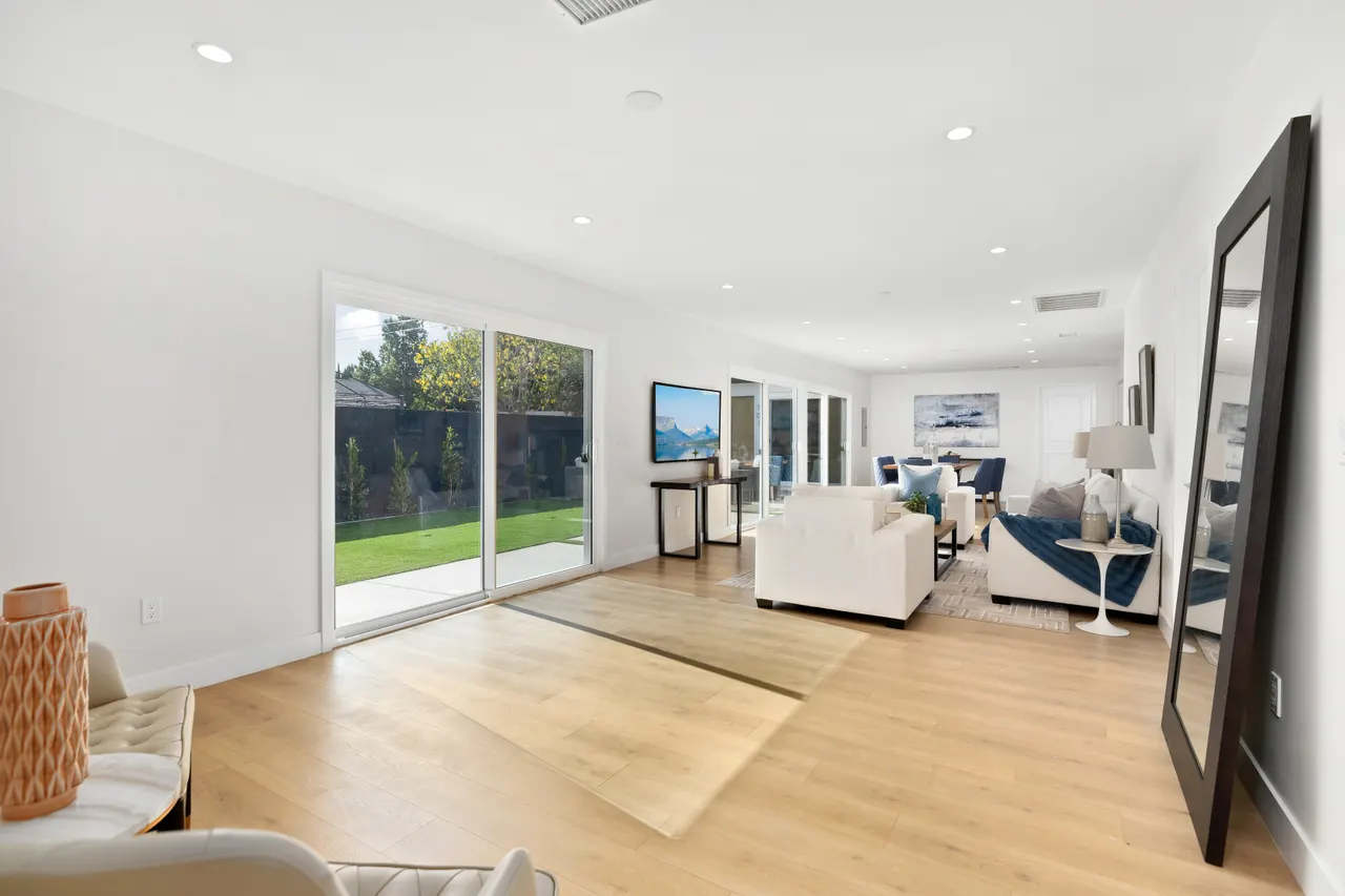 Open great room with wide-plank light wood floors, a white sofa, a floor mirror leaning against the wall, and floor-to-ceiling sliding glass doors to the backyard