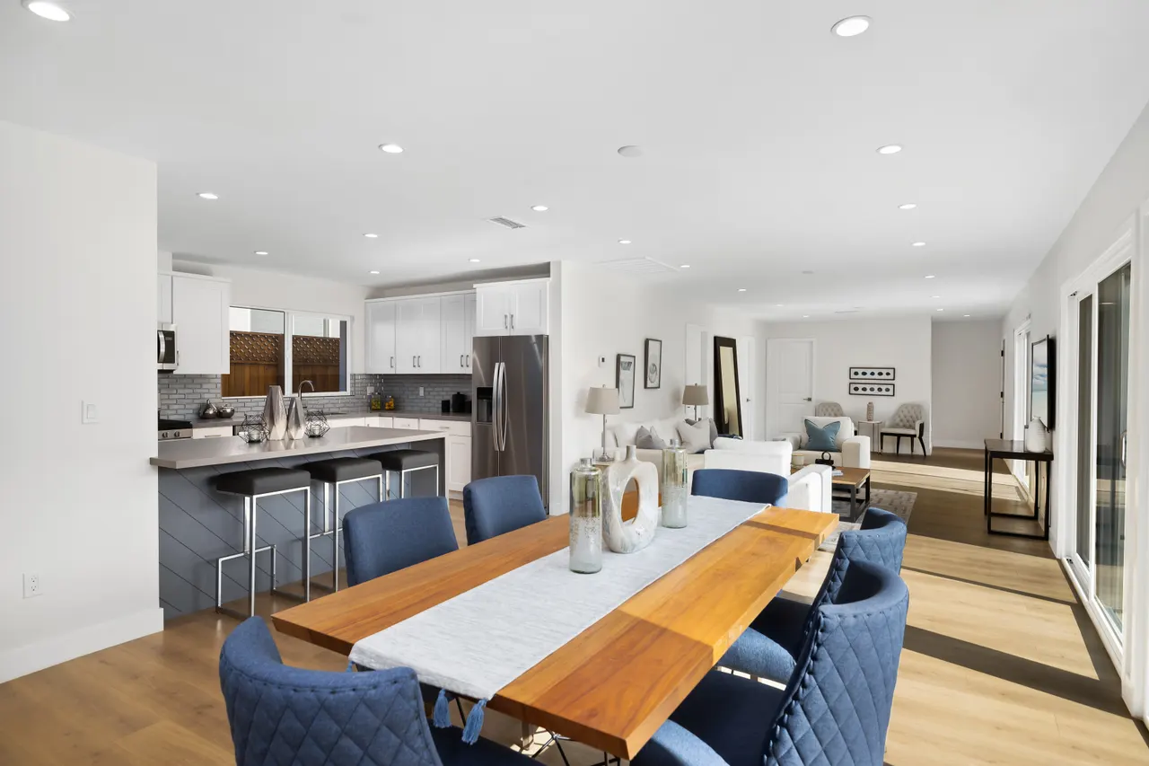 Open-concept kitchen, dining, and living area in one sightline, with an island bar, a long dining table with navy upholstered chairs, and sliding glass doors to the yard
