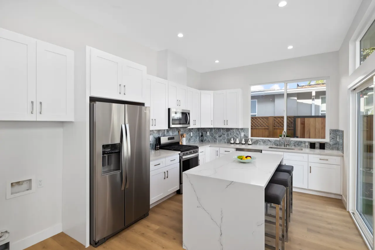 Kitchen with white shaker cabinets, a waterfall quartz island with pendant lighting, a stainless steel refrigerator and range, and a window over the sink