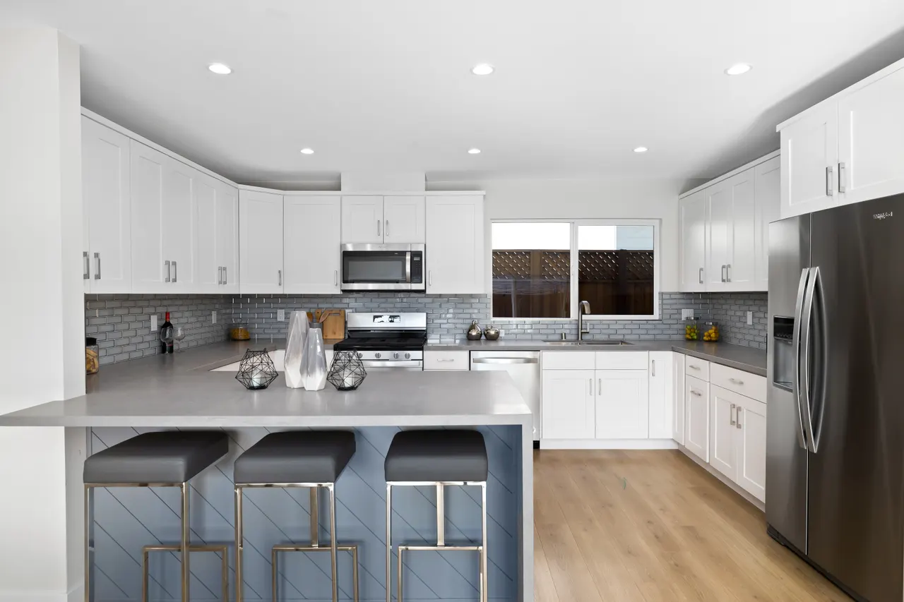 Kitchen close-up with white cabinetry, gray subway tile backsplash, gray quartz counters, stainless appliances, and an island with black bar stools