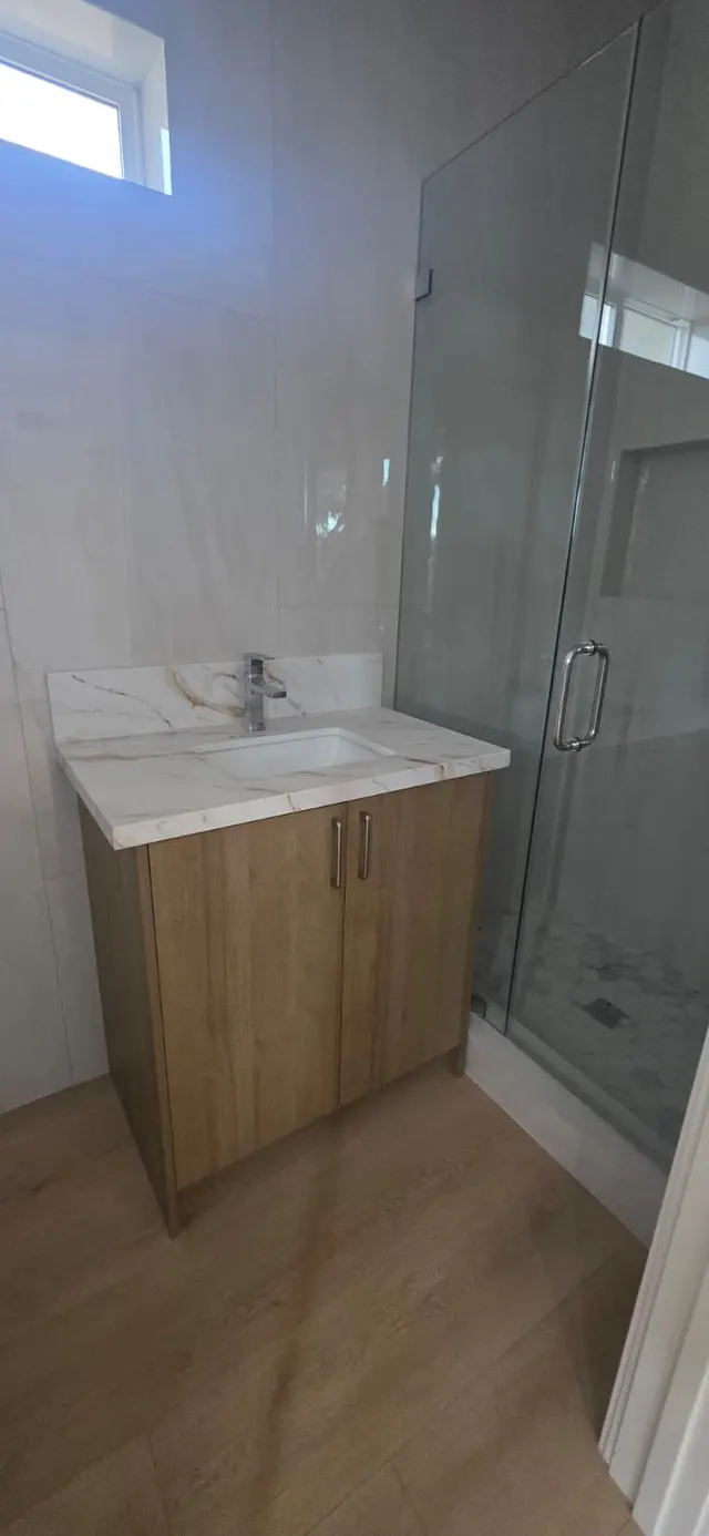 Bathroom with a wood-tone vanity, marble-look countertop, and a frameless glass shower