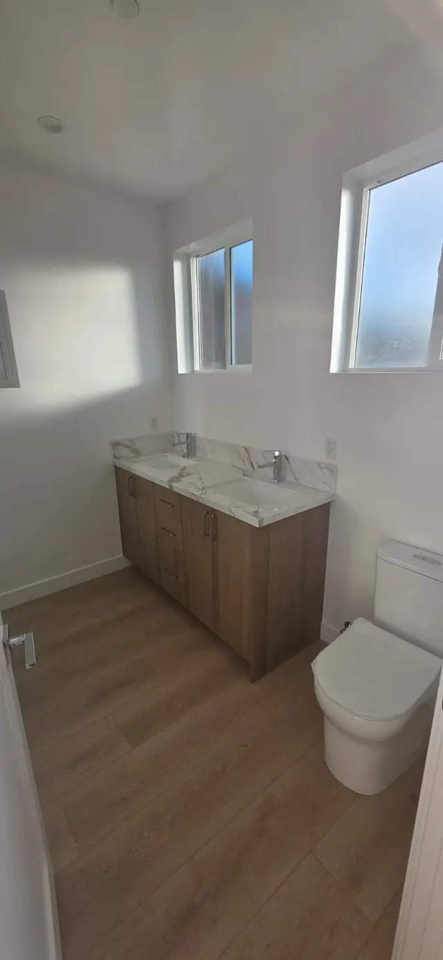 Bathroom with a wood-tone double vanity, marble-look countertop, toilet, and two windows