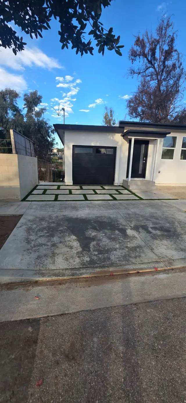 Single-story home exterior with a black garage door, navy front door, and a paver-and-turf walkway under a palm tree