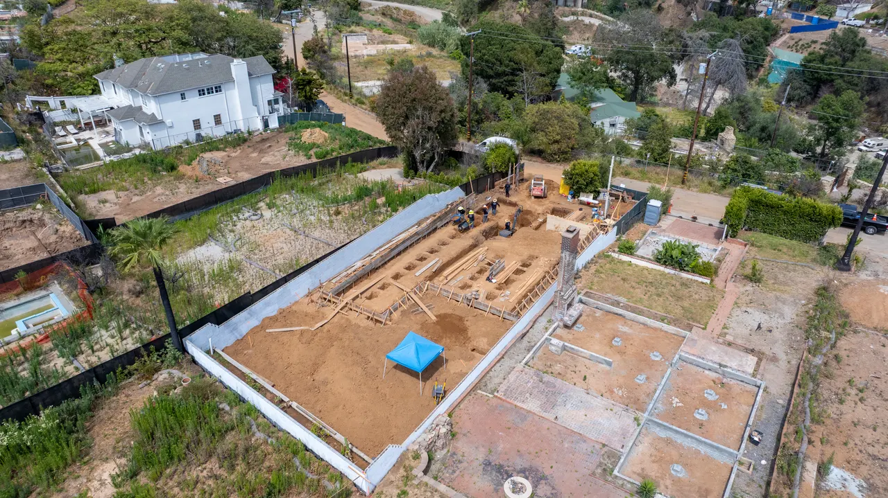 Aerial view of the full property during foundation work, with the neighboring two-story home and street visible beyond the perimeter fencing