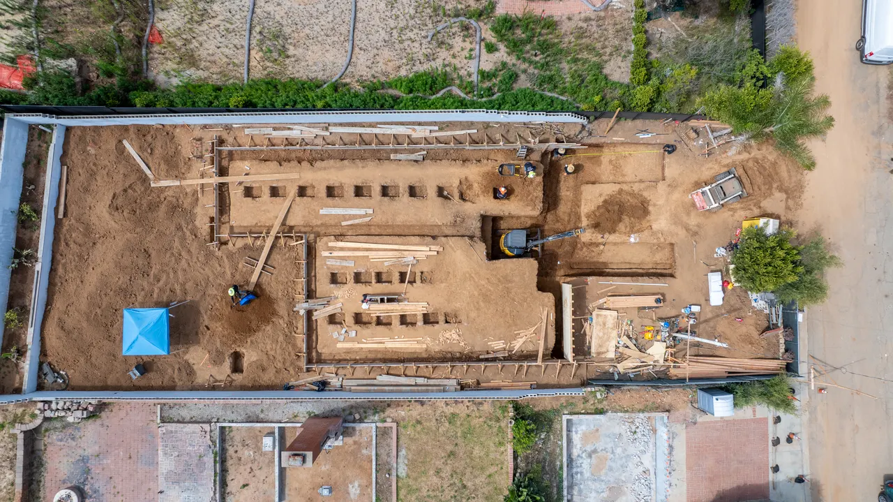 Direct overhead aerial of the foundation footprint showing footing trenches, formwork boards, a concrete pump, lumber staged for framing, and crew on site