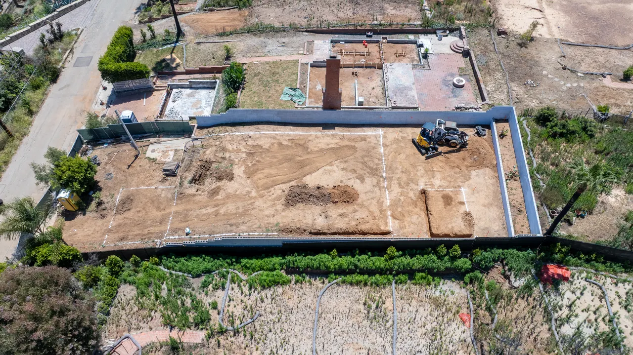 Aerial close-up of the freshly poured perimeter foundation walls with a graded dirt pad and mini skid steer on site