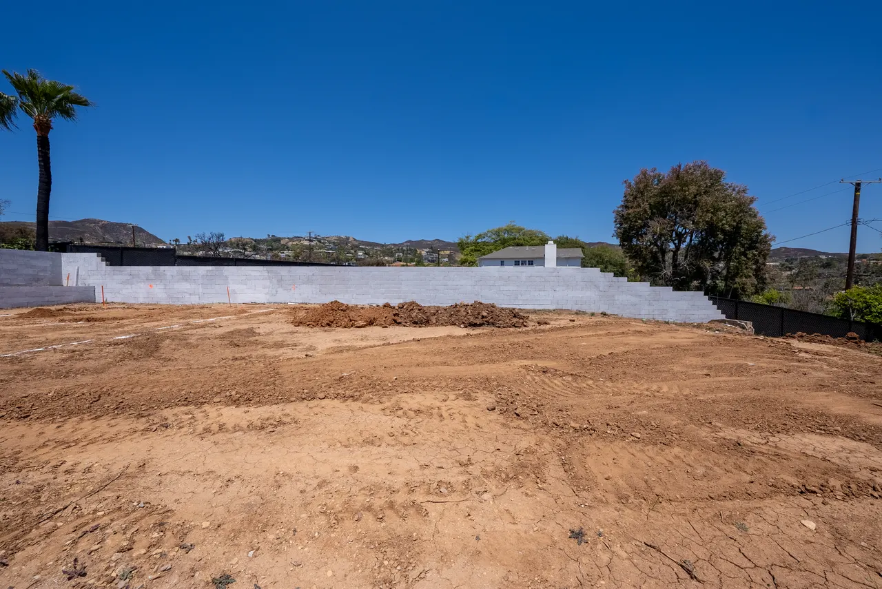 Ground-level view of the finished concrete block retaining wall bordering the graded building pad, with hills and rooftops beyond