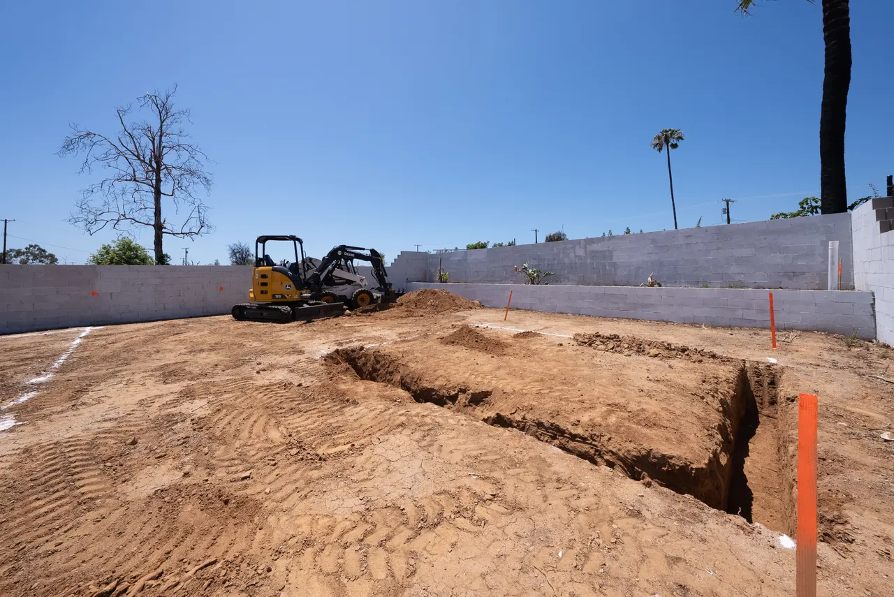 Ground-level view of a mini excavator digging a footing trench along the foundation line, with the concrete block retaining wall in the background