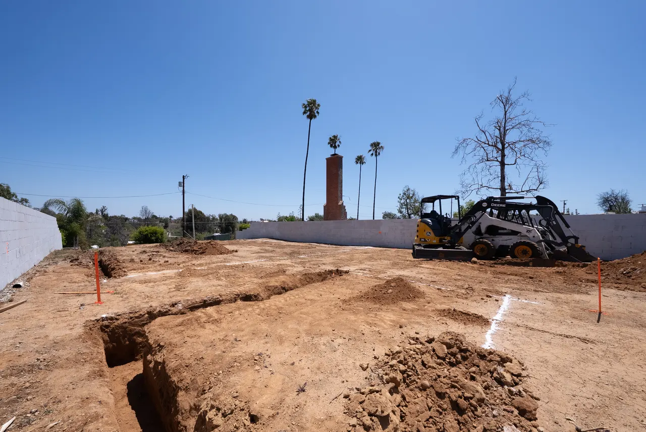 Ground-level view of footing trenches being excavated with a skid steer, palm trees and a brick chimney in the background