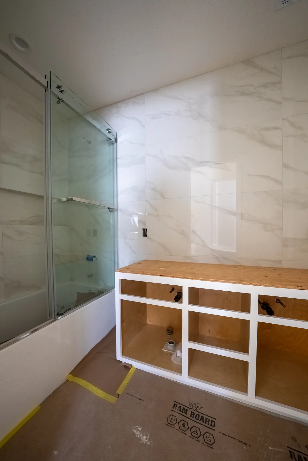 Bathroom with a frameless glass shower enclosure and marble-look tile walls; the vanity is unfinished plywood awaiting doors and countertop