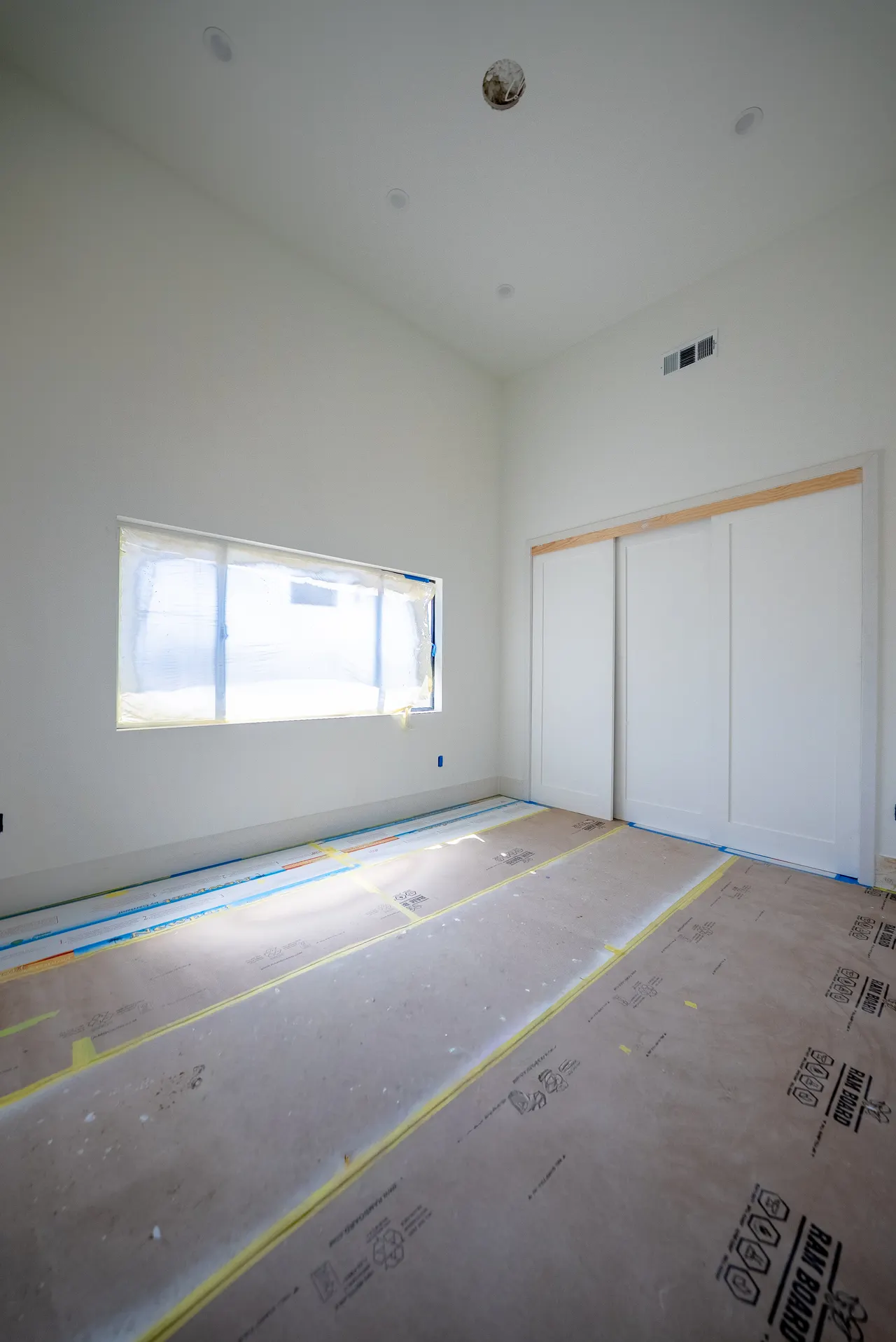 Bedroom with white paneled sliding closet doors and a window; floor is paper-protected and window film is still in place