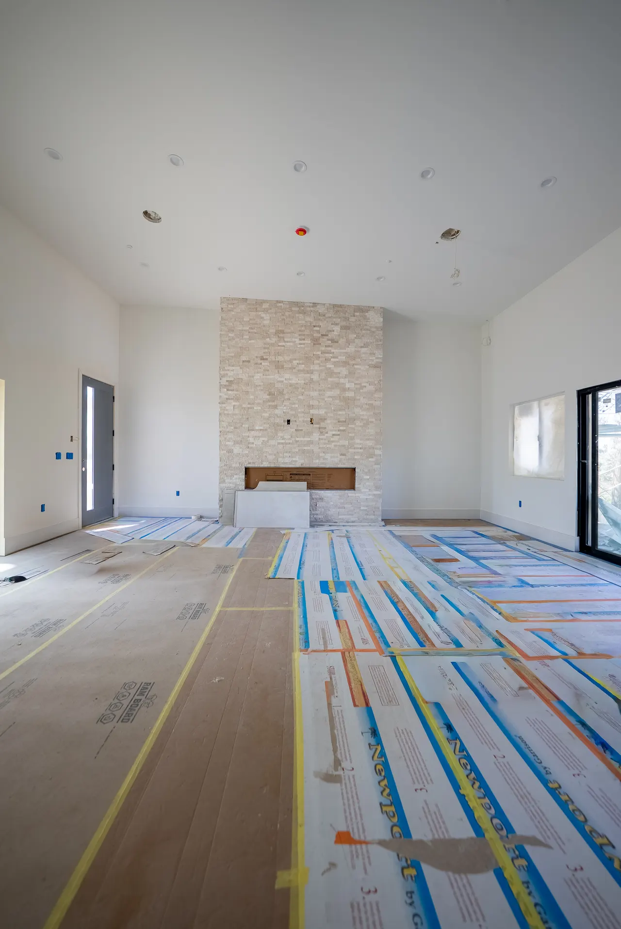 Living room with the stacked-stone fireplace wall, front entry door, and large windows; hardwood floor is paper-protected