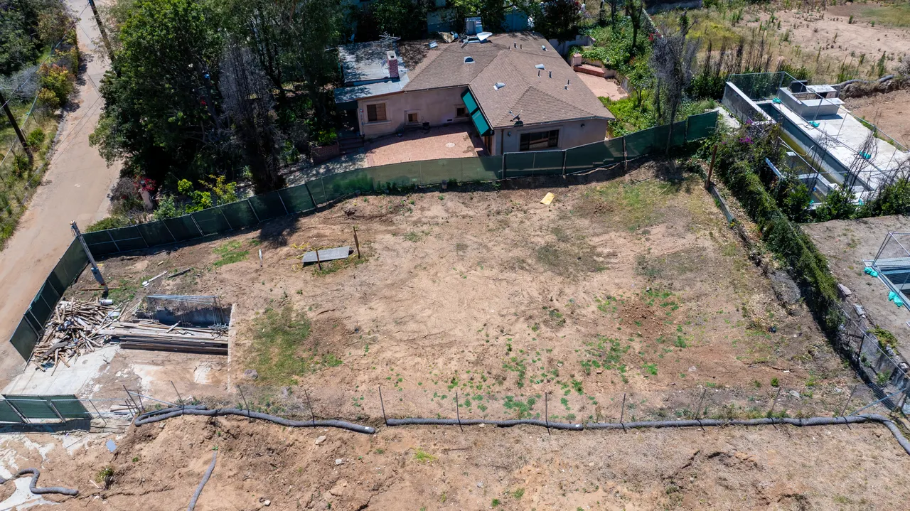 Aerial view of the graded dirt building lot enclosed by perimeter fencing, with a neighboring pink house and pool visible along the property line