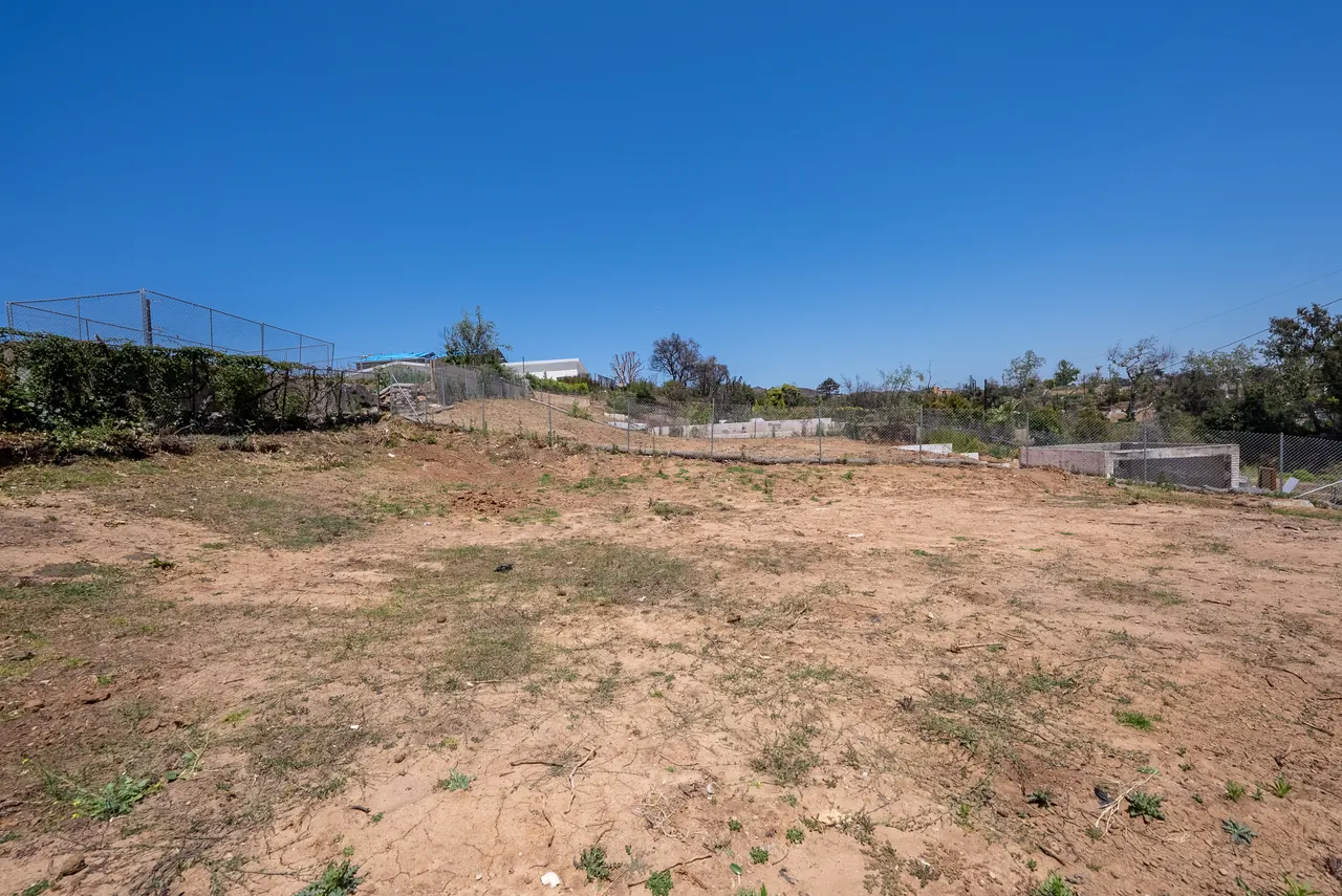 Ground-level view across the open, graded lot toward surrounding trees and hillside under a clear blue sky