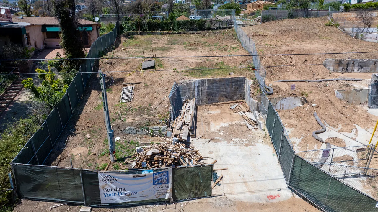 Aerial view of the foundation excavation area enclosed by green construction fencing, with excavated soil and building materials staged on site