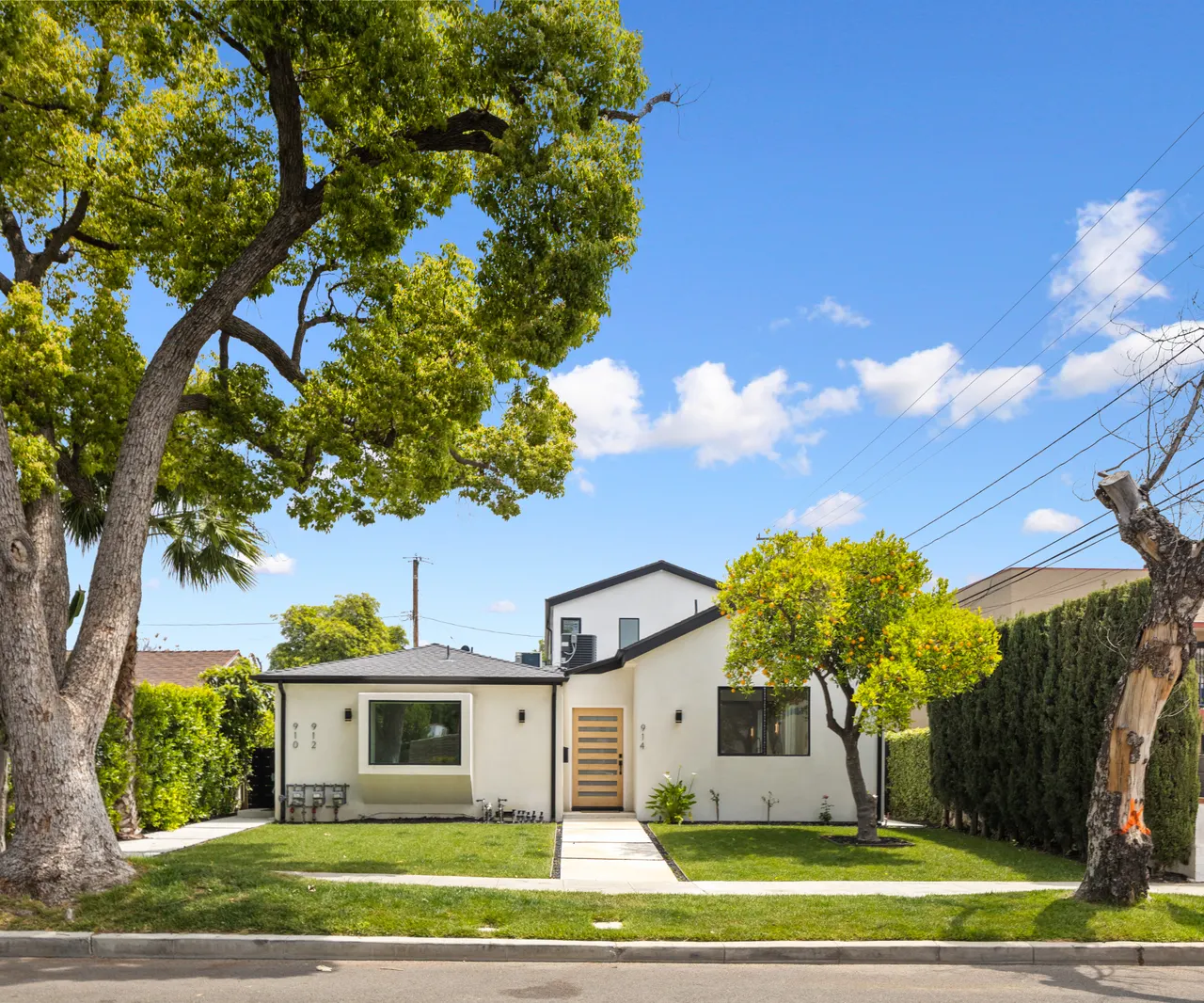 Front exterior of a white stucco custom home with black trim, a wood slat entry door, and a lush green lawn shaded by a mature street tree