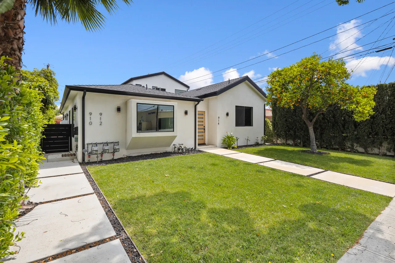 Front elevation of the modern home with a wood slat entry door, black-framed windows, a two-story addition behind, and a fruit-bearing orange tree in the yard