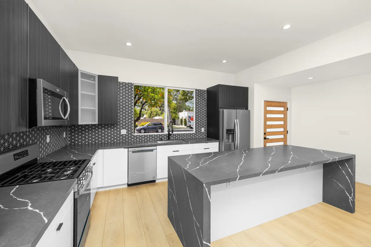 Kitchen with a dark waterfall-edge island, charcoal wood-grain cabinetry, patterned tile backsplash, and stainless steel appliances