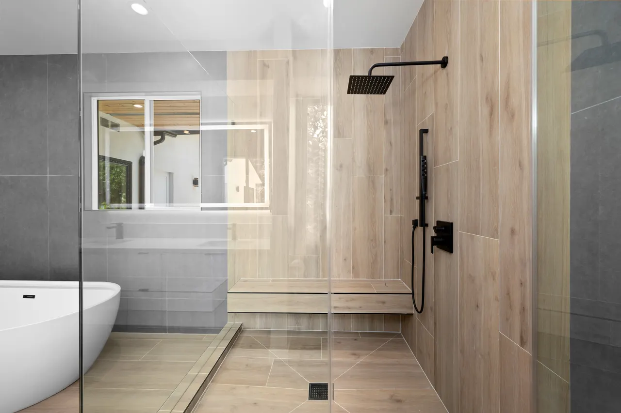 Walk-in shower detail with a matte black rain shower head and handheld sprayer against wood-look tile walls and a built-in bench