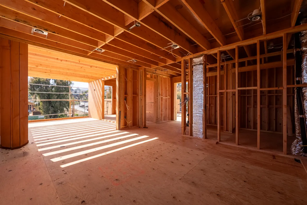 Framed interior opening onto a covered outdoor deck, with late-day light streaking across the subfloor and hillside views beyond