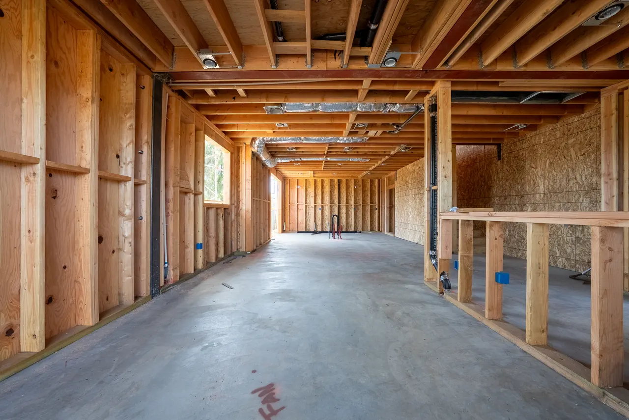 Long framed corridor marked for the ADU, with stacked lumber, wrapped ductwork, and rough plumbing overhead