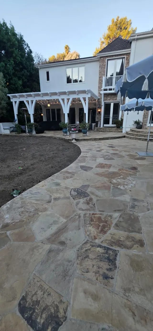 Wide dusk view of the two-story house with a string-lit pergola, flagstone patio, and blue patio umbrellas