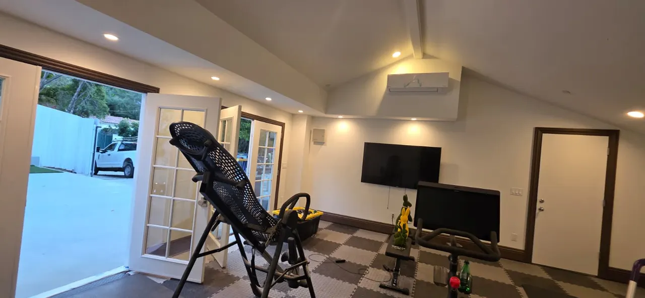 Home gym with a vaulted ceiling, two mounted TVs, an inversion table and exercise bike on black-and-gray checkered rubber flooring, and open French doors to the patio