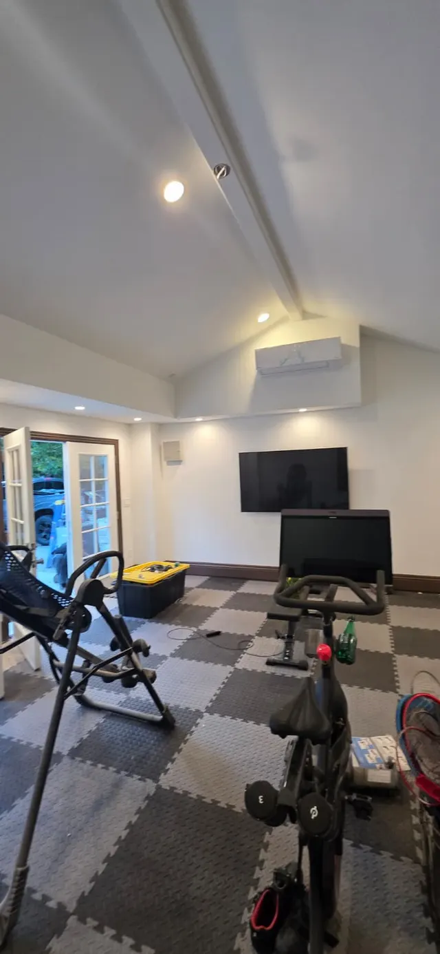 Home gym corner with a mounted TV, wall-mounted mini-split unit, inversion table, and exercise bike on checkered rubber flooring