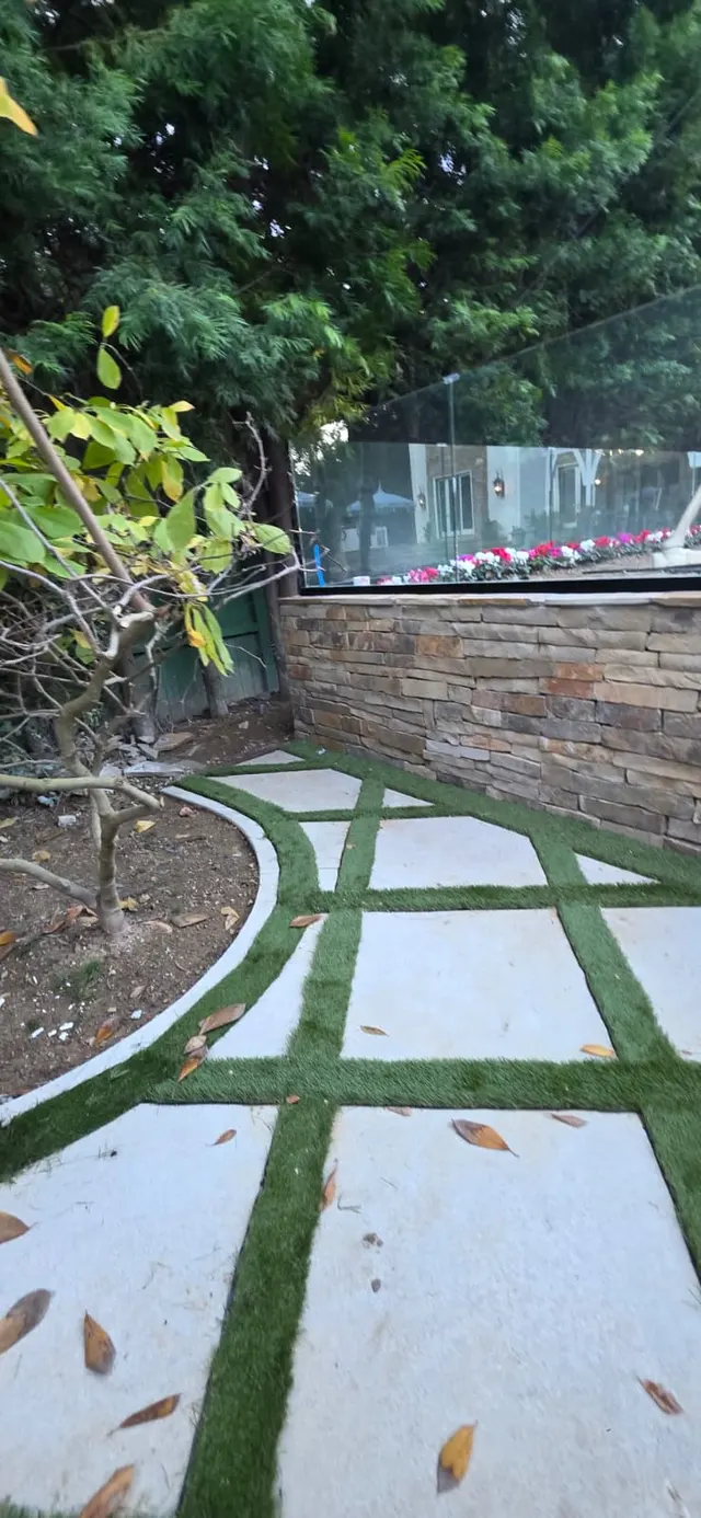 Turf-and-concrete checkerboard paving curving around a young tree, next to a stacked stone retaining wall topped with a glass railing and flowering plants beyond