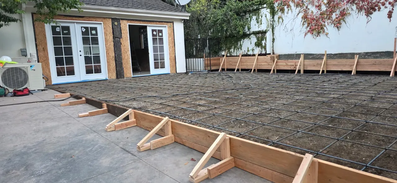 Rebar grid and wood forms set for a concrete slab pour behind a house, with French doors and wood sheathing visible on the building