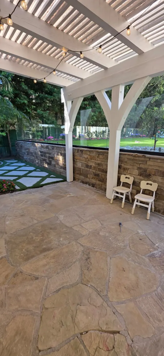 Flagstone patio under a white wood pergola strung with string lights, bordered by a stacked stone retaining wall with glass railing and a turf-and-paver walkway