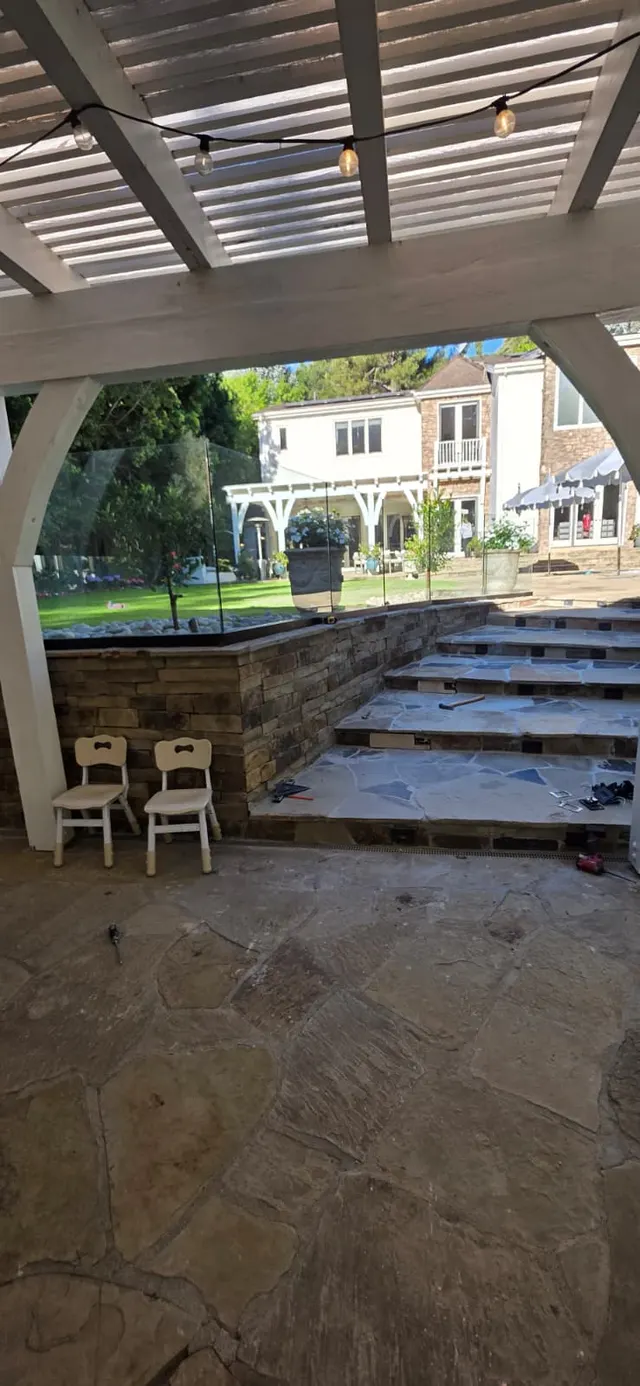 View from beneath one pergola across a green lawn to a second white pergola, with a stacked stone retaining wall, uplit flagstone stairs, and glass railing between them